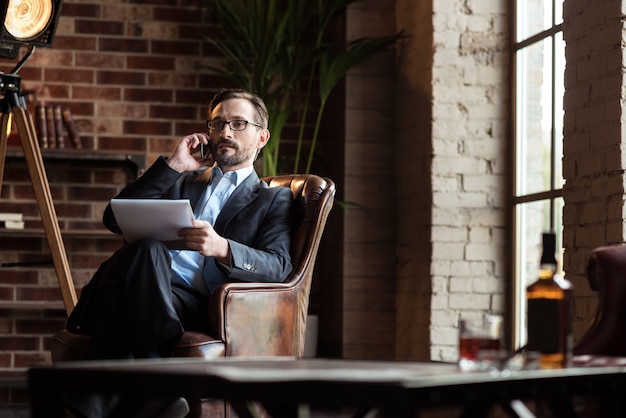 Arbeitsgespräch. Hübscher stilvoller ruhiger Geschäftsmann, der einen teuren Anzug trägt und während der Arbeit in seinem Arbeitszimmer auf dem Handy spricht