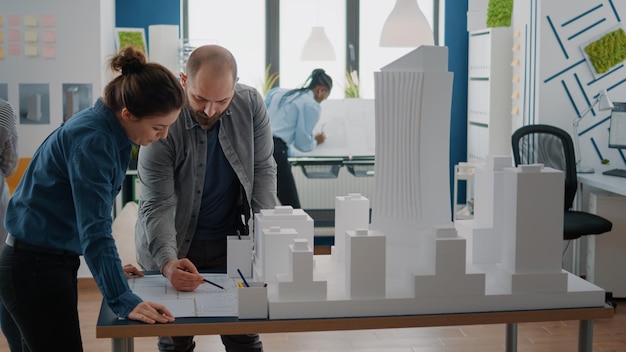 Foto arbeitskollegen, die maquette und blaupausen analysieren, planen auf dem tisch, an architektur und gebäudemodell zu arbeiten. mann und frau, die zusammenarbeiten, um baulayout und strategie im büro zu entwerfen.