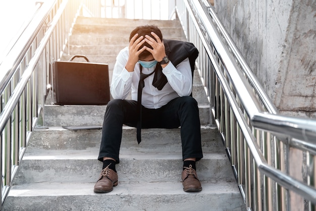 Arbeitslose betonten den jungen asiatischen Geschäftsmann im Anzug, der das Gesicht mit den Händen bedeckt. Ausfall- und Entlassungskonzept für die Krankheit Covid-19, Arbeit und Stress. Geschäftsleute verzweifeln niedrige Wirtschaftskrise