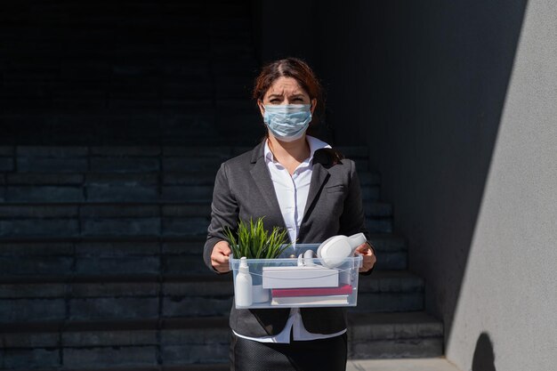 Arbeitslose Frau mit Schutzmaske steht mit einer Kiste persönlicher Gegenstände auf der Treppe. Geschäftsdame wird entlassen. Die globale Wirtschaftskrise führt zu einem Arbeitsausfall aufgrund der Insolvenz des Unternehmens