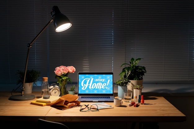 Arbeitsplatz am Fenster im dunklen Raum mit Laptop, Papiersack mit Croissant, Blumen, Getränken, Kosmetikartikeln, Büchern auf dem Tisch und Lampe oben