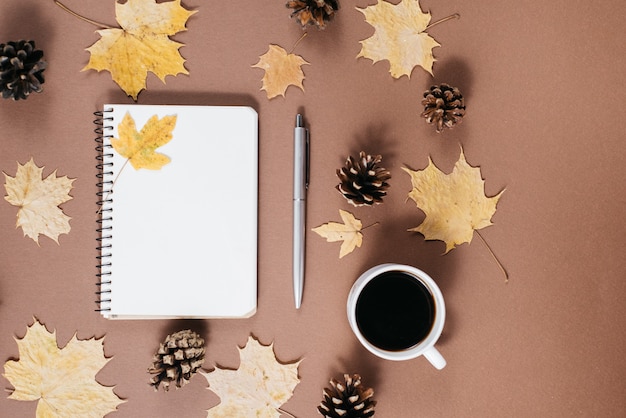 Arbeitsplatz mit goldenen Ahornblättern, Kaffeetasse, Stößen, Notizbuch und Stift auf braunem Hintergrund.