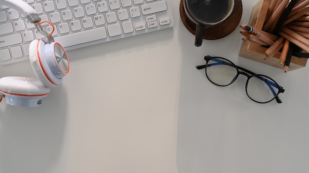 Arbeitsplatz mit kopfhörer, kaffeetasse, tastatur, brille und kopierraum auf weißem tisch.