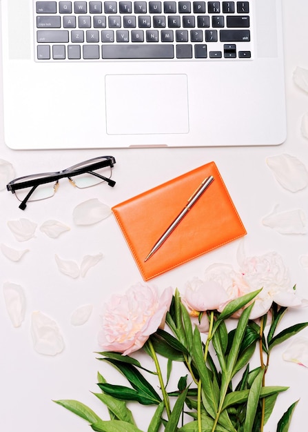Arbeitsplatz mit Laptop, Brille, Notizbuch, Stift und Pfingstrosenblumen auf dem weißen Tischhintergrund. Flache Lage, Draufsicht