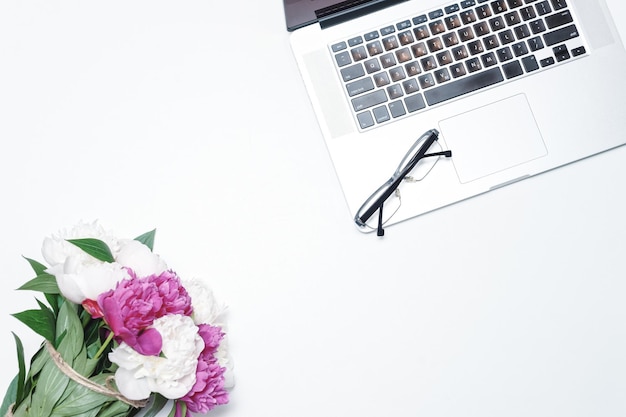 Arbeitsplatz mit Laptop, Brille und mit rosa und weißen Pfingstrosen auf dem weißen Tischhintergrund. Flache Lage, Draufsicht