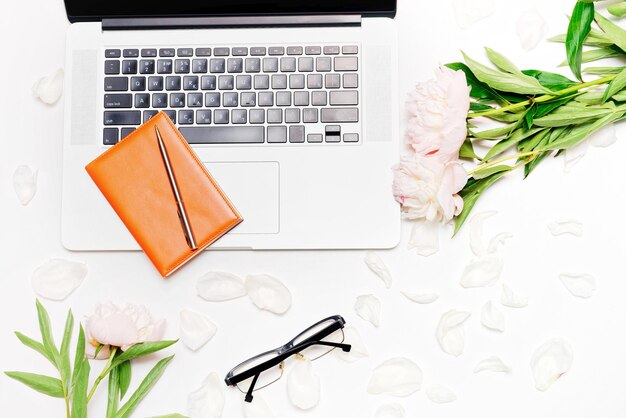 Arbeitsplatz mit Laptop, Notizbuch, Stift, Brille und Pfingstrosenblumen auf dem weißen Tischhintergrund. Flache Lage, Draufsicht