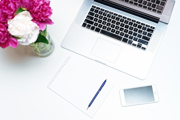 Foto arbeitsplatz mit laptop, notizbuch, stift und rosa und weißen pfingstrosenblumen auf dem weißen tischhintergrund. flache lage, draufsicht