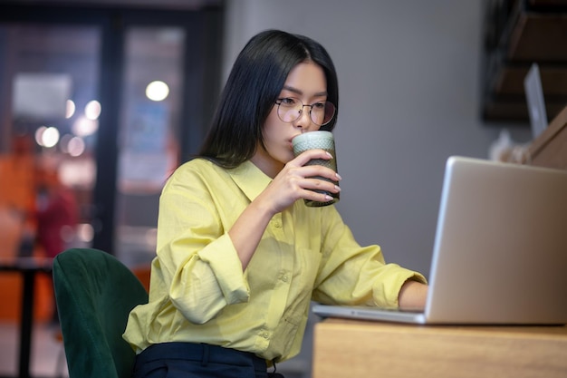 Arbeitsplatz. Nette asiatische junge Frau in den Brillen am Laptop, der Kaffee trinkt