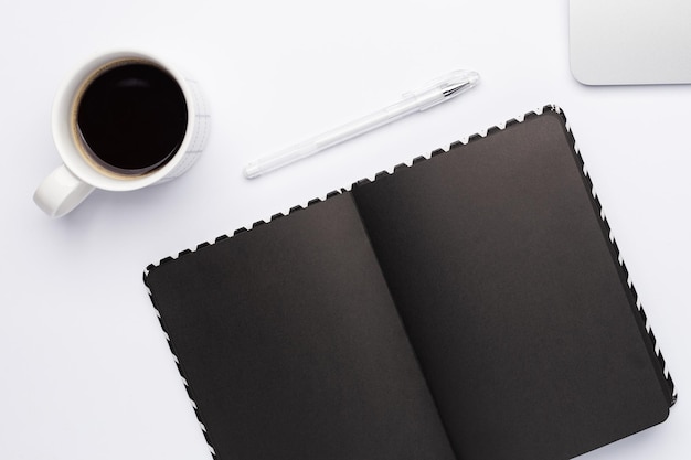 Arbeitsplatz zur Quarantänezeit, leeres dunkles Notizbuch mit Tasse Kaffee auf weißem Tisch. Weißer Farbarbeitsplatz mit Laptop, Minimalismus.