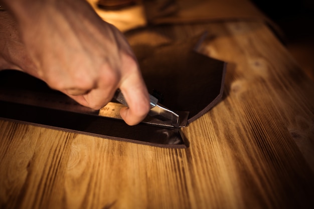 Arbeitsprozess des Ledergürtels in der Lederwerkstatt. Mann, der Handwerkszeug hält und arbeitet. Gerber in alter Gerberei. Holztabellenhintergrund. Nahaufnahme Mann Arm