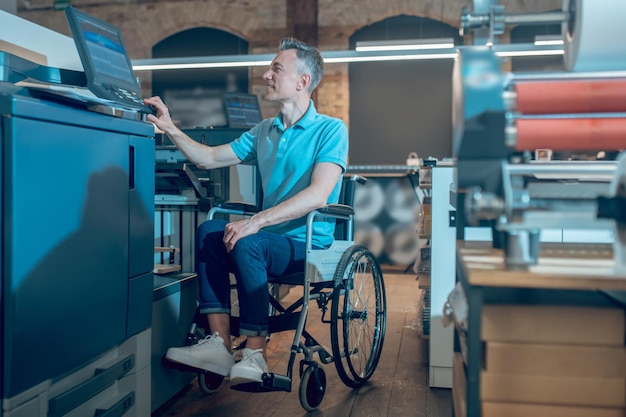 Arbeitsprozess. Lächelnder Mann im Profil auf Rollstuhl, der das Bedienfeld der Büroausstattung in geräumigen beleuchteten Büroräumen berührt