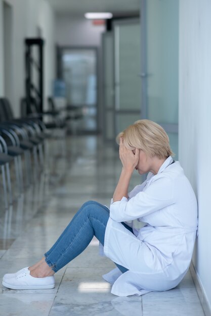 Arbeitsüberbelastung. Müde blonde Ärztin sitzt auf dem Boden und bedeckt ihr Gesicht