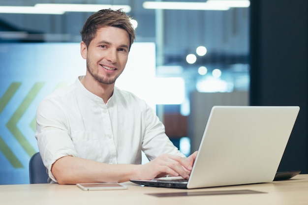 Arbeitsumfeld Porträt eines jungen gutaussehenden männlichen Geschäftsmannes, der an einem Schreibtisch im Büro sitzt und an einem Laptop-Tablet-Handy arbeitet Gewinne arbeitet Er schaut in die Kamera und lächelt
