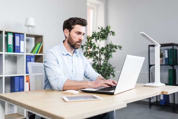 Arbeitsumgebung im Büro Porträt eines jungen männlichen Wirtschaftswissenschaftlers, der im Büro am Schreibtisch auf einem Laptop arbeitet und mit Papierdokumenten arbeitet