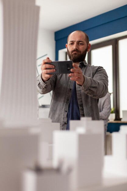 Architekt, der ein Smartphone verwendet, um ein Video des architektonischen Modells eines Wolkenkratzers aufzunehmen, um es in sozialen Medien zu veröffentlichen. Bärtiger Ingenieur fotografiert 3D-Modell aus weißem Schaumstoff mit Smartphone im modernen Büro.