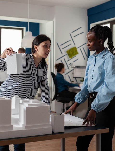 Architekt, der einem Ingenieurmitarbeiter, der neben dem Wohnprojekt-Maquette in einem modernen Architekturbüro steht, ein Gebäudemodell im Schaummaßstab präsentiert. Team von Architekten, die kreatives Denken tun.