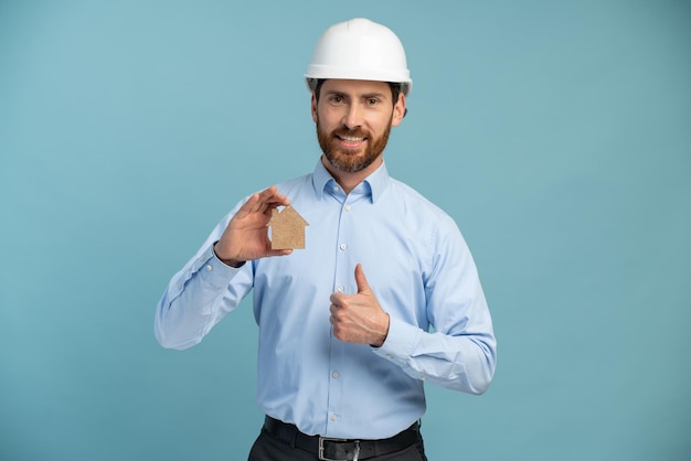 Architekt Mann mit lächelndem Gesicht im Bauarbeiterschutzhelm, der in die Kamera blickt und eine hausförmige Figur über isoliertem Hintergrund hält Ingenieurkonzept