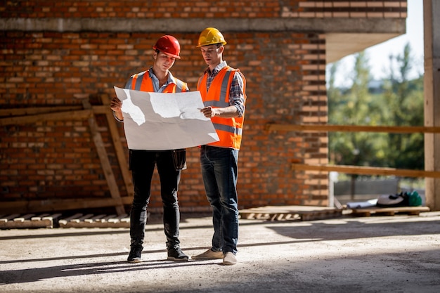 Architekt und Bauleiter in orangefarbenen Arbeitswesten und Helmen diskutieren die Dokumentation auf einem Backsteinmauerhintergrund.