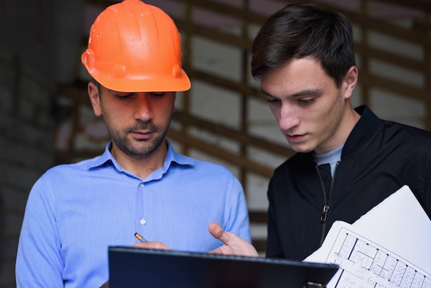 Architekteningenieur, der orange Sturzhelm am Arbeitsstandort erklärt dem Kunden die Projektpläne trägt