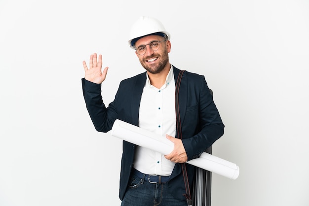 Architektenmann mit Helm und Halteplänen lokalisiert auf weißer Wand, die mit Hand mit glücklichem Ausdruck salutiert