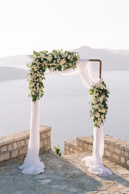 Un arco de boda rectangular cubierto con una tela blanca se encuentra en una plataforma de observación sobre el mar