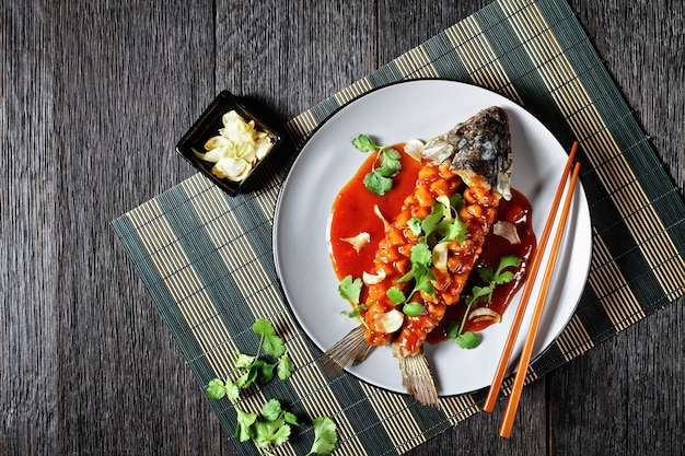 Ardilla carpa: pescado agridulce chino con cilantro fresco y ajo seco encima servido en una placa gris sobre una mesa de madera vieja oscura, vista superior, primer plano