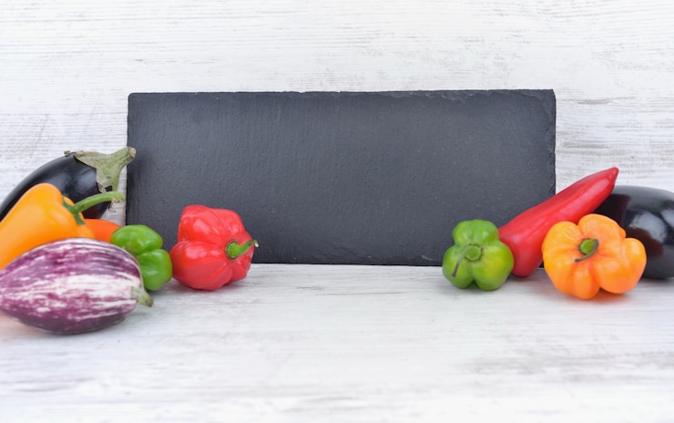 Foto ardósia preta com pimentos coloridos e berinjela de cada lado em uma mesa branca