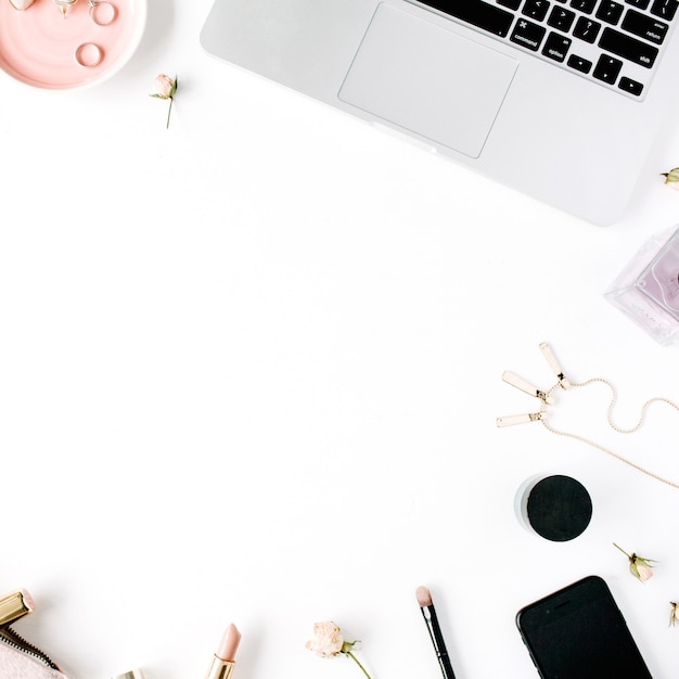 área de trabalho da mesa feminina com laptop, embreagem, cosméticos, telefone, óculos de sol, botões de rosa de batom em branco.