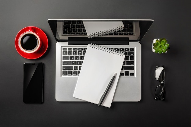 Área de trabalho de escritório preta elegante com laptop, notebook, xícara de café vermelha e telefone celular. vista do topo.