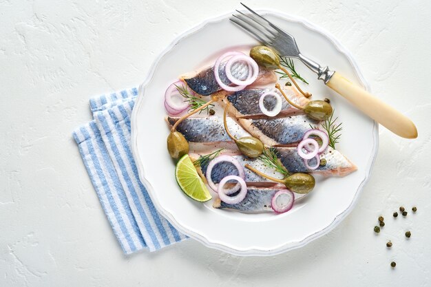 Arenque salado con especias, hierbas y cebolla en un plato blanco sobre fondo de piedra clara