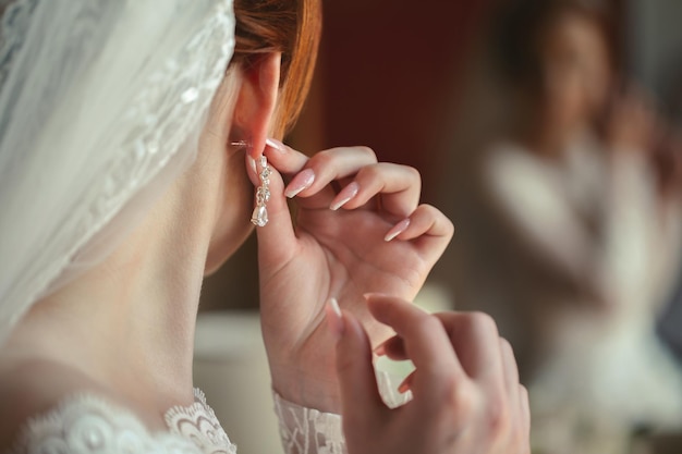 Aretes de boda en la mano de una mujer, ella lleva aretes, honorarios de novia, novia de la mañana, vestido blanco, aretes