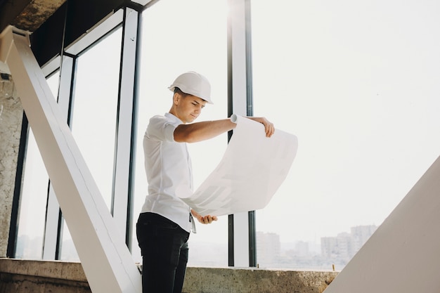 Arhitect adulto seguro que mira el plan del edificio en construcción cerca de una ventana grande.