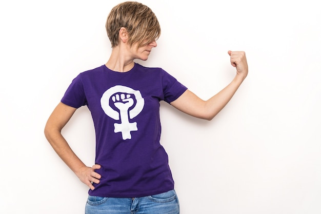 Arm der blonden Frau, die ein lila T-Shirt mit dem Symbol des internationalen feministischen arbeitenden Frauentags auf einer weißen Wand schwingt