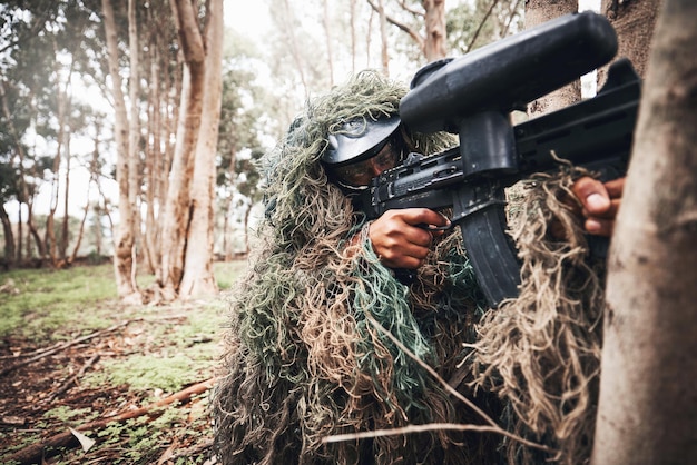 Arma de paintball forrest e homem com mira em árvores para estratégia de jogo de guerra ao ar livre ou foco em camuflagem natural Atirador de elite e capacete para segurança com os olhos no alvo para vencer em jogos militares