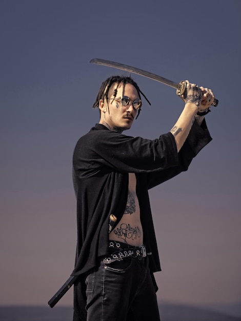 Arma samurai y japón. Concepto de artes marciales. Guerrero con rastas y ropa abierta mostrando el torso tatuado. Concentración y zen. Hombre con espada katana de pie en el cielo azul.