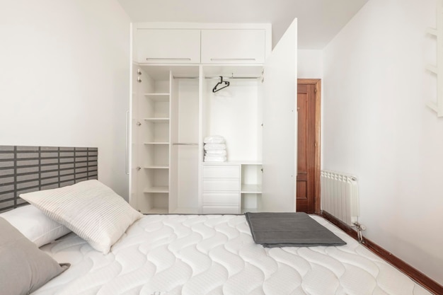 Armario blanco abierto con estantes, cajones, baúl de ropa y perchas negras en el dormitorio con colchón kingsize y radiador de aluminio