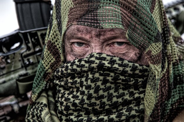 Armeeschütze, Airsoftspieler in Tarnuniform und Ladungsträger, Abdeckcape auf dem Kopf, bewaffnetes Dienstgewehr mit optischem Visier, Gesicht mit Shemagh verstecken, auf Feld stehen, in die Ferne schauen