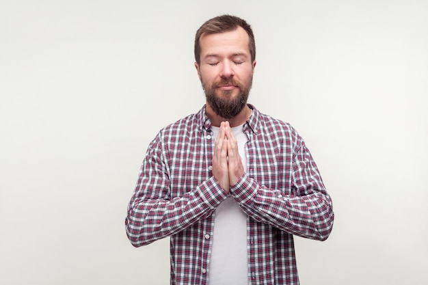 Armonía y equilibrio. Retrato de un hombre barbudo con camisa a cuadros meditando con los ojos cerrados y la cara tranquila, tomándose de la mano en gesto de namaste de oración, mente pacífica. Foto de estudio aislado sobre fondo blanco.