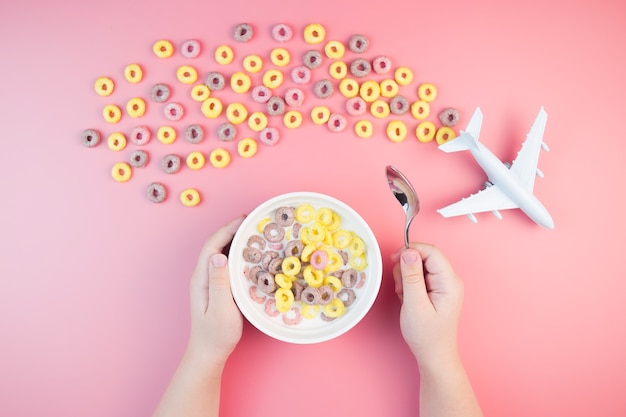 Aros de maíz de frutas con leche en el recipiente, manos de los niños, avión en rosa