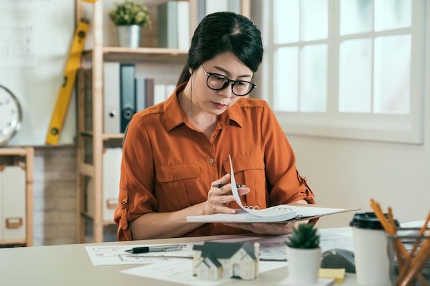 arquitecta asiática atractiva trabajando en bocetos de planos en la oficina. una joven ingeniera en prácticas leyó una nota en un cuaderno con cara seria. mujer elegante diseñadora de interiores pensamiento creativo concentrado