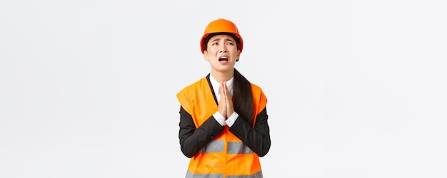 Arquitecta gerente de construcción asiática desesperada e imprudente suplicando a Dios usando casco de seguridad tomándose de la mano en oración suplicando pidiendo ayuda a Dios rogando sobre fondo blanco