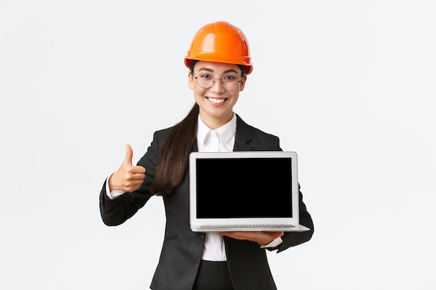 Arquitecta profesional sonriente, gerente de construcción en la fábrica que muestra el gráfico, diagrama positivo, hace el pulgar hacia arriba en señal de aprobación y sostiene la pantalla de la computadora portátil, usa casco de seguridad