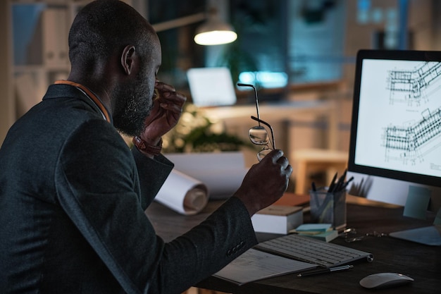 Arquitecto africano cansado sentado en la mesa frente al monitor de la computadora con plano mientras trabaja en la oficina hasta la noche