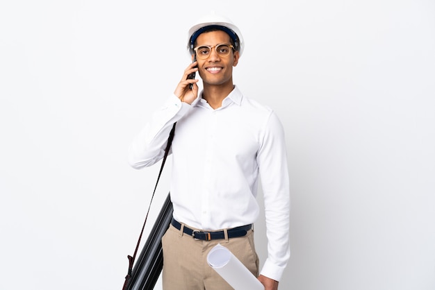 Arquitecto afroamericano hombre con casco y sosteniendo planos sobre pared blanca aislada _ manteniendo una conversación con el teléfono móvil con alguien