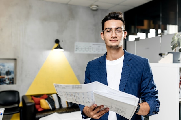 Arquitecto elegante vestido con chaqueta a cuadros azul y jeans trabaja con planos en la oficina moderna.