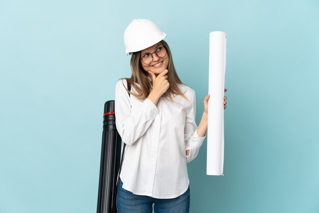 Arquitecto eslovaco chica sosteniendo planos aislado sobre fondo azul mirando hacia el lado y sonriendo