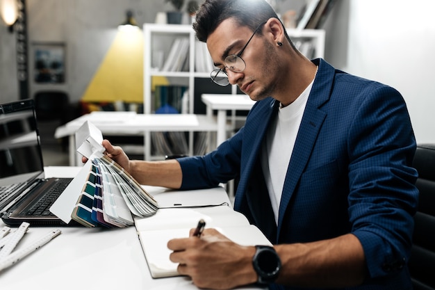 El arquitecto con gafas y chaqueta azul está trabajando con un catálogo de colores en el escritorio de la oficina.