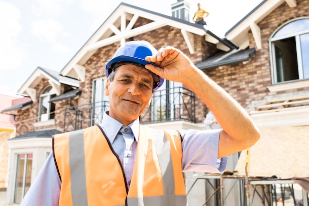 Arquitecto hombre indio feliz en el sitio de construcción mirando a la cámara gerente de construcción confiado con casco azul y chaleco de seguridad amarillo retrato de ingeniero civil maduro exitoso