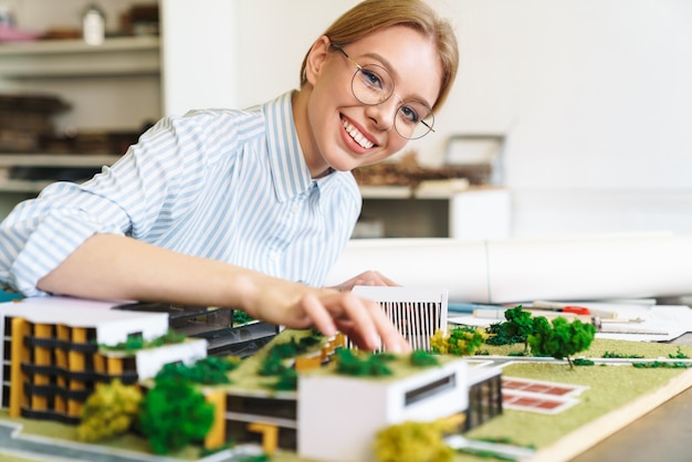 Arquitecto joven alegre en anteojos diseñando proyecto con modelo de casa y sentado en el lugar de trabajo