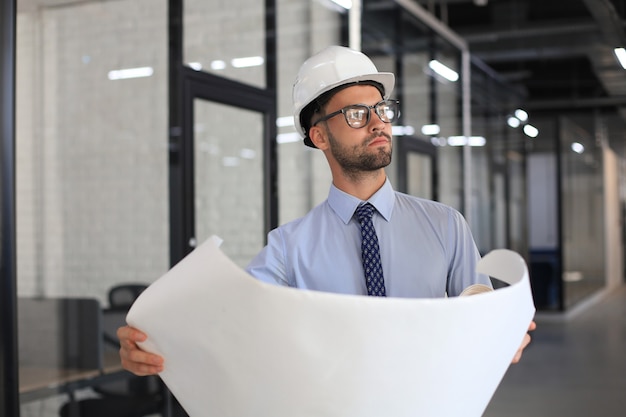 Arquitecto joven confiado en casco sosteniendo un plano y mirando el área de trabajo.